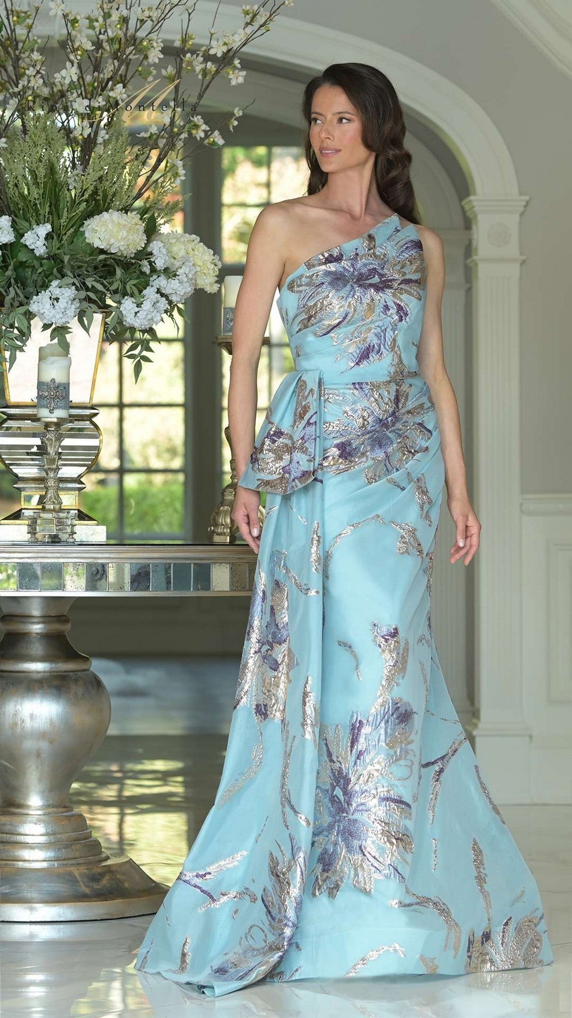 A woman in the Rina Di Montella RD3233 long formal dress in Tiffany Multi stands indoors near a tall floral arrangement and a silver table.